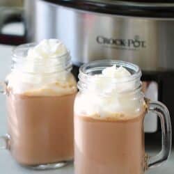 Two mugs of hot chocolate topped with whipped cream are in front of a Crock-Pot on a countertop.