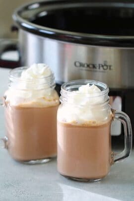 Two mugs of hot chocolate topped with whipped cream are in front of a Crock-Pot on a countertop.