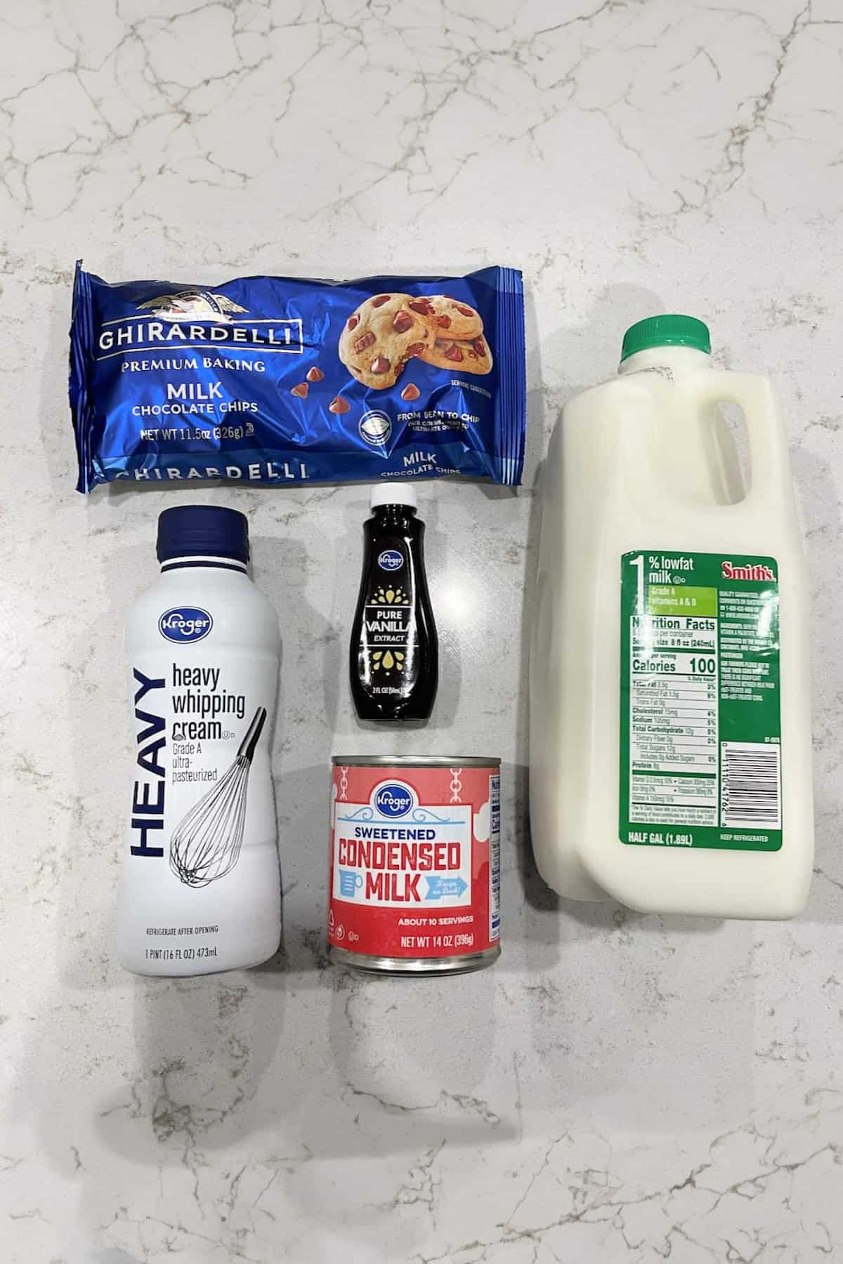 A selection of ingredients on a countertop: a bag of milk chocolate chips, a bottle of heavy whipping cream, a can of sweetened condensed milk, chocolate syrup, and a gallon of 1% milk.