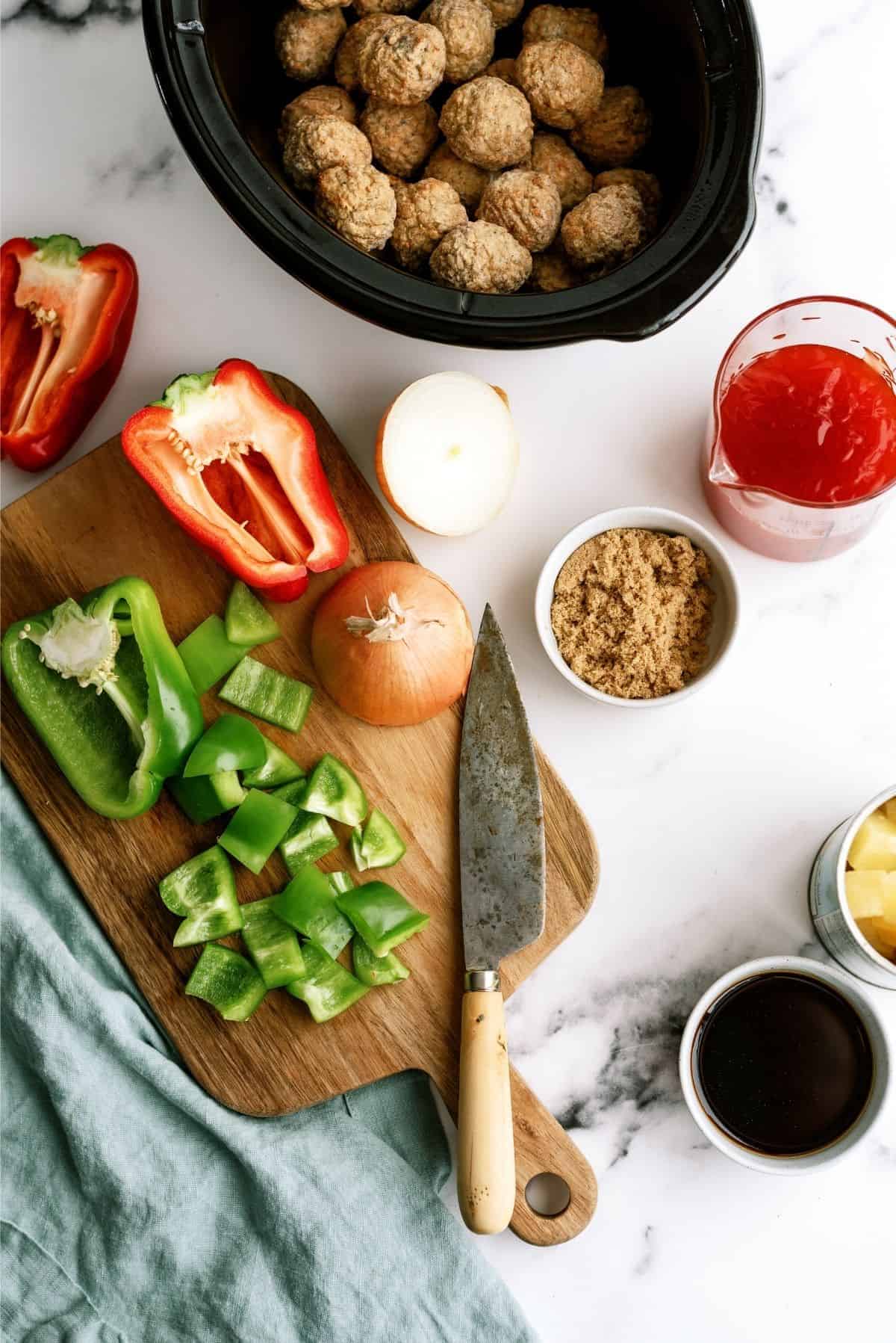 A countertop with chopped green and red bell peppers, an onion, a knife on a cutting board, and various ingredients including meatballs in a slow cooker, brown sugar, soy sauce, and a red sauce.