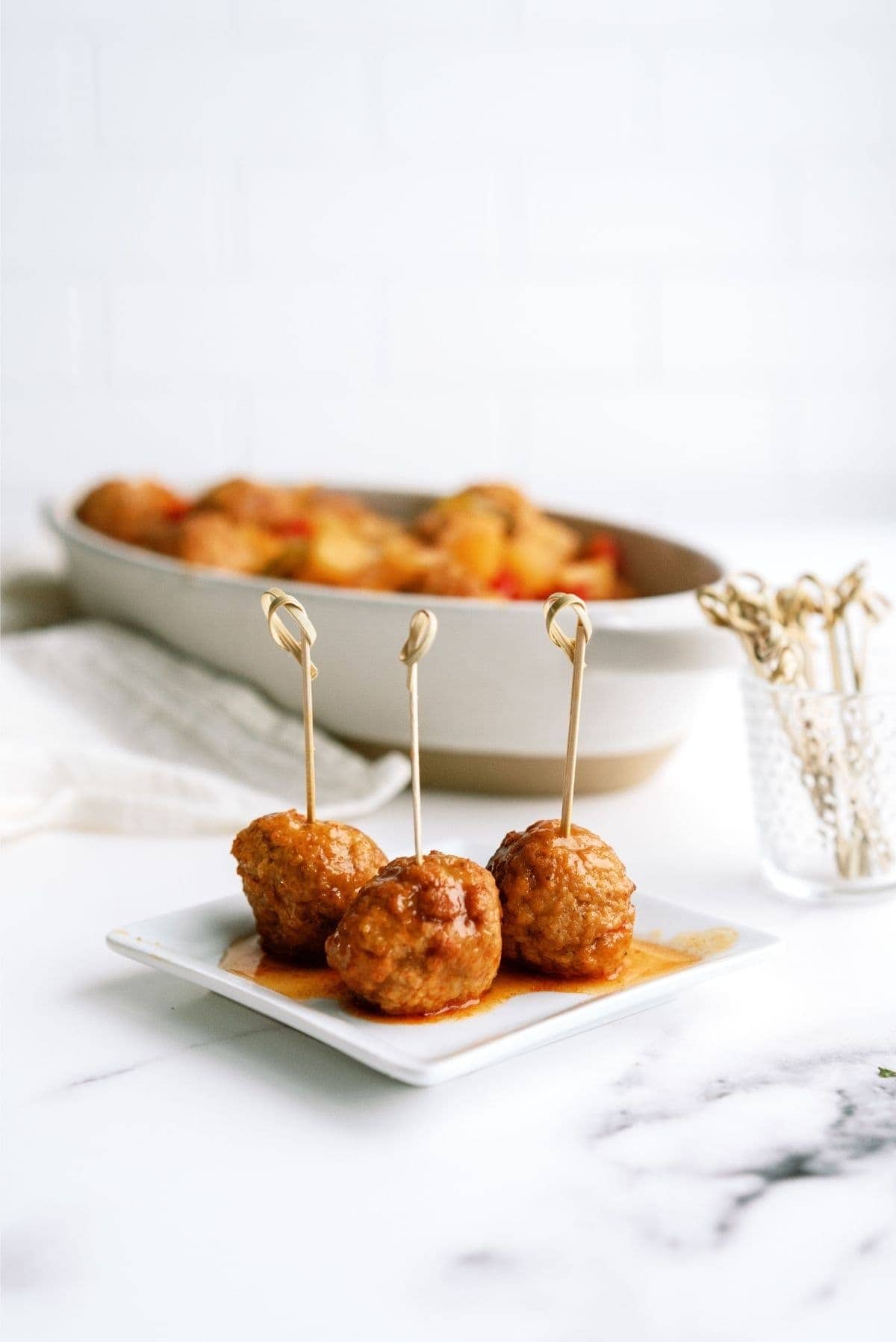 Three sauced meatballs on skewers are presented on a small white plate, with a dish holding more meatballs blurred in the background. A jar of additional skewers sits beside the dish.