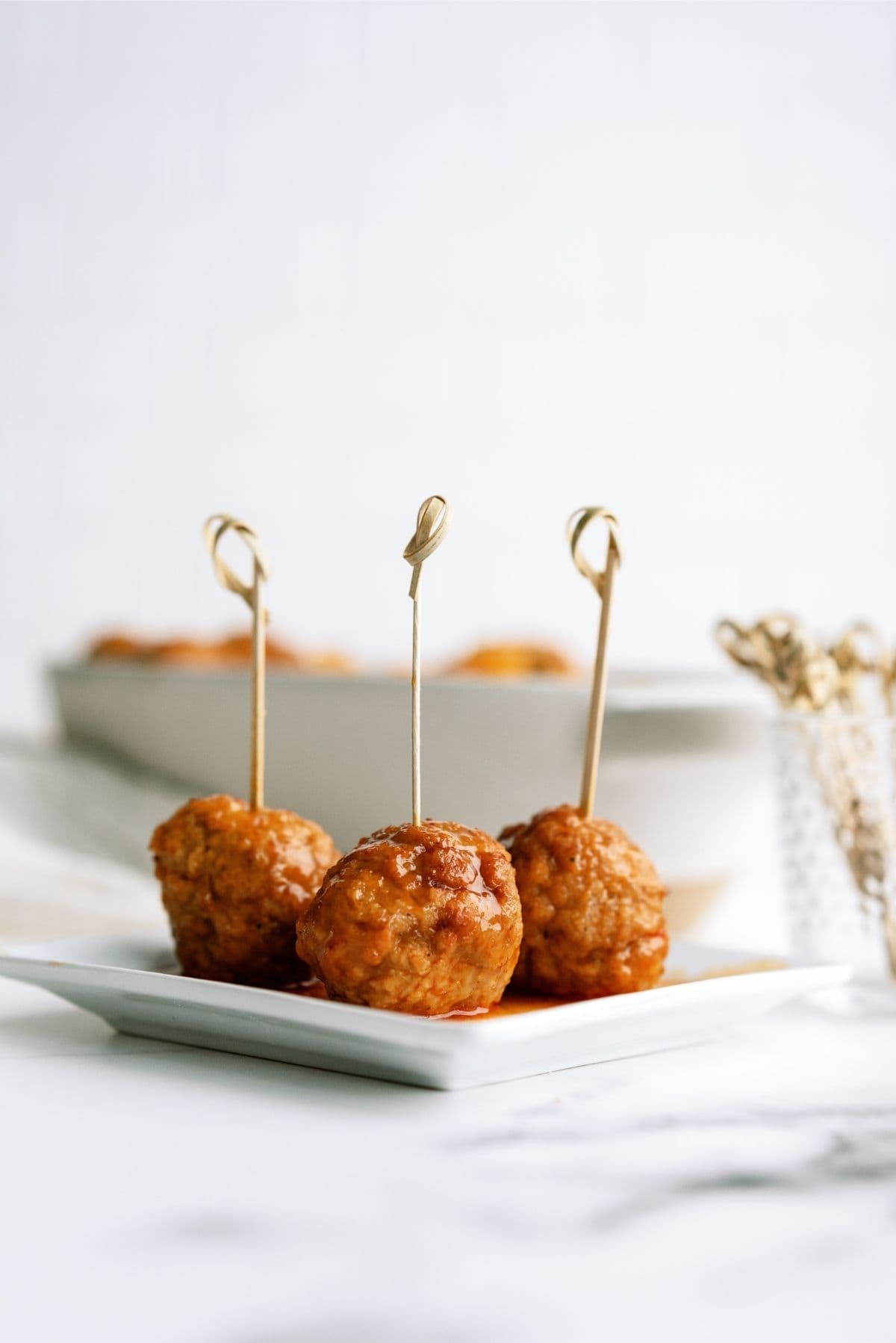 Three meatballs on skewers are arranged on a small white plate, with a dish containing more meatballs slightly out of focus in the background.