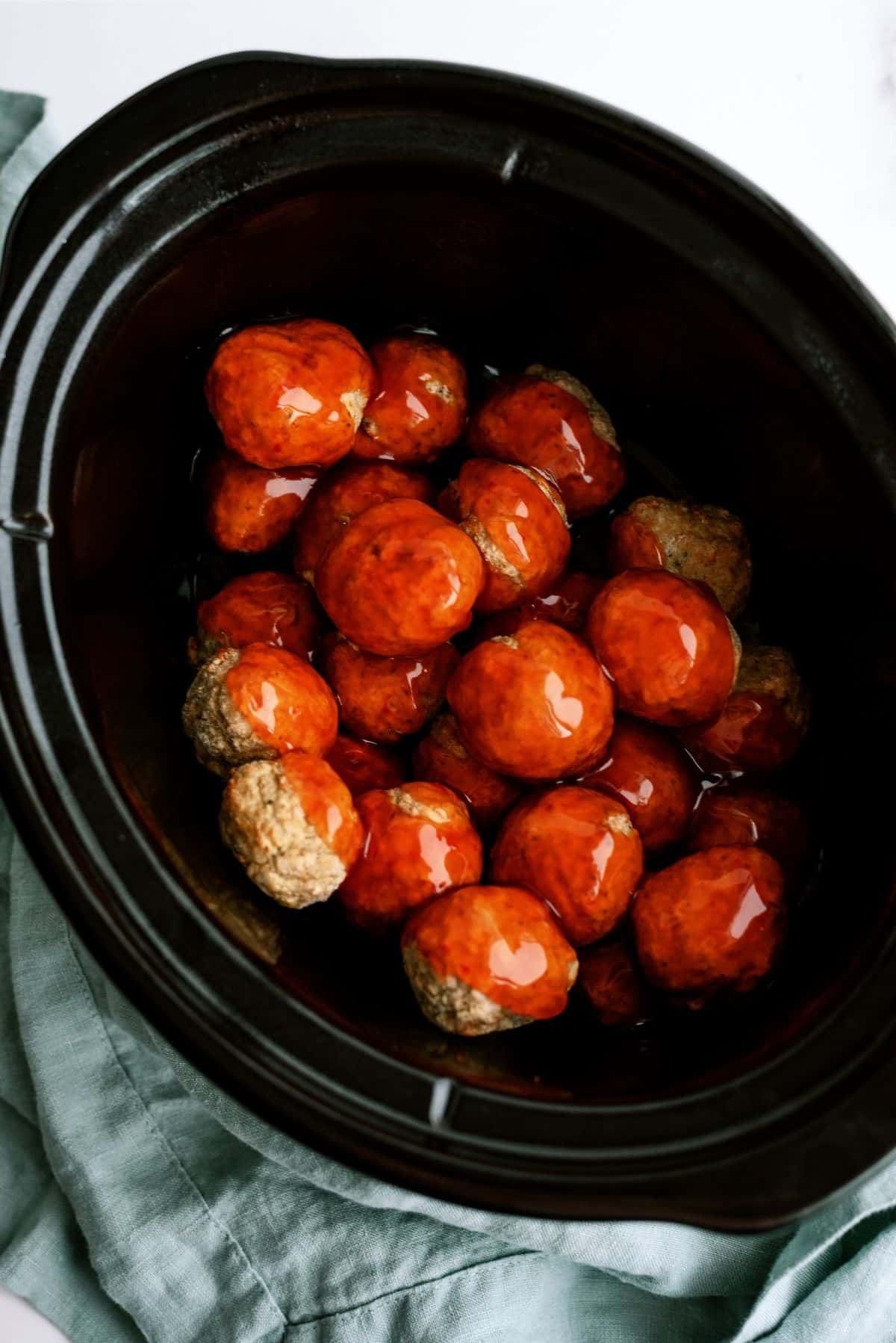 Slow cooker filled with glazed meatballs placed on a light green cloth.