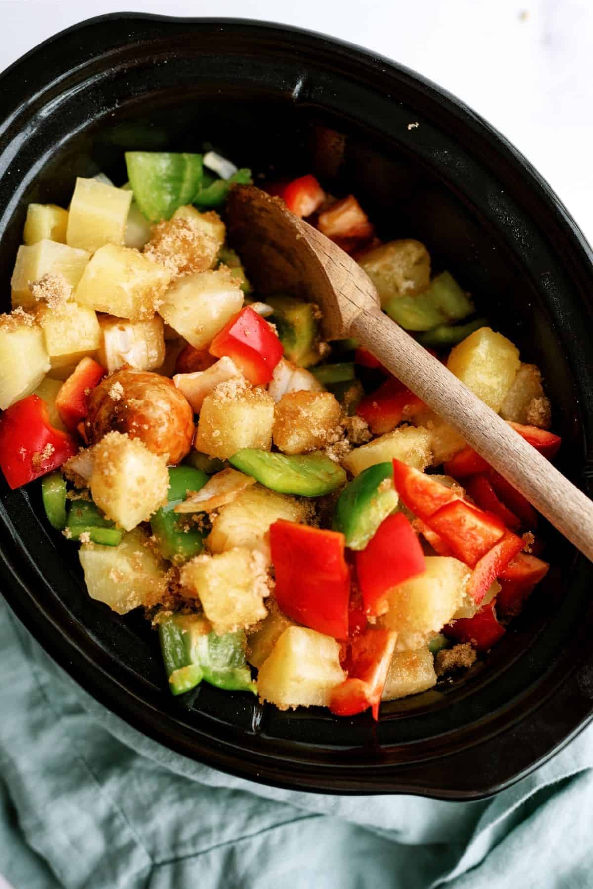 A slow cooker filled with chopped green and red bell peppers, pineapple chunks, white onions, and brown sugar, with a wooden spoon. A gray cloth is partially visible under the cooker.