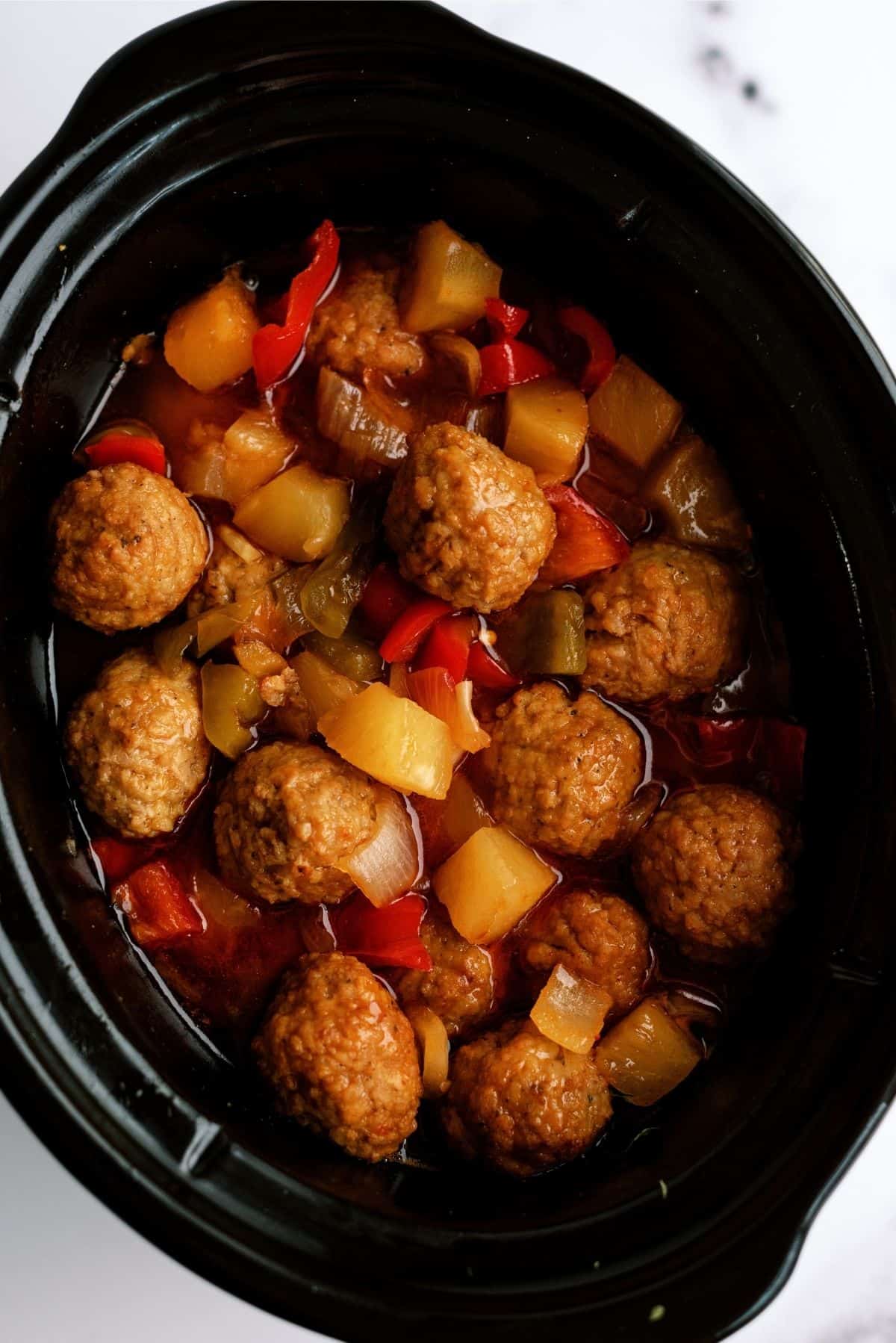 A black slow cooker filled with meatballs, pineapple chunks, and assorted vegetables in a red sauce.