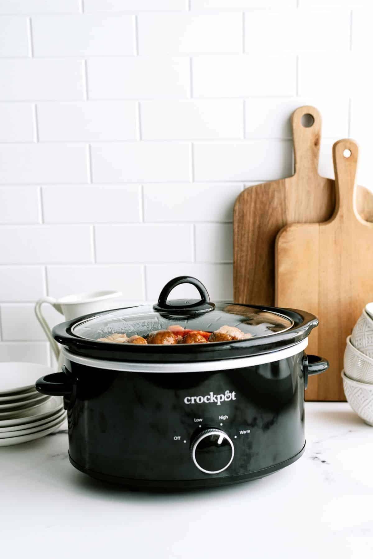 A black Crockpot filled with food sits on a white kitchen countertop amidst stacked plates, a pitcher, and wooden cutting boards.