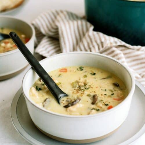 A bowl of creamy soup with vegetables and mushrooms, served with a black spoon inside. A striped cloth is in the background.