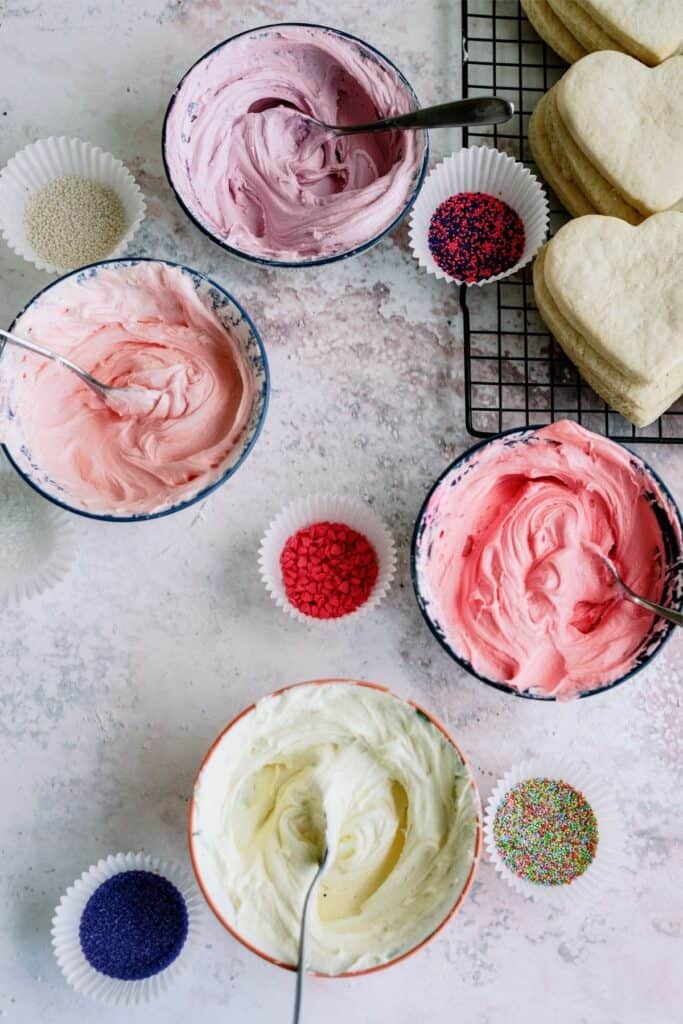 4 bowls of different colored frosting, little bowls of sprinkles and heart shaped sugar cookies on a cooling rack.