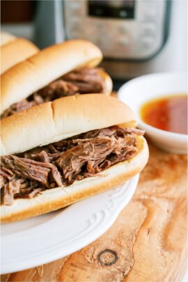 Two Instant Pot French Dip Sandwiches on a white plate with a small bowl of au ju sauce off to the side and an Instant Pot in the background.