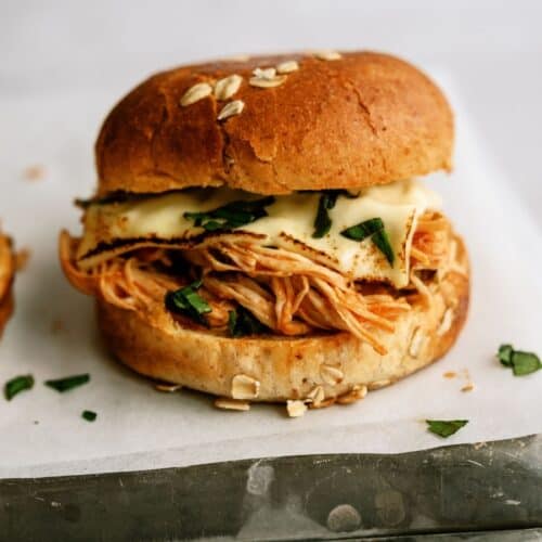 A sandwich with shredded chicken, melted cheese, and greens in a sesame seed bun, placed on white parchment paper.