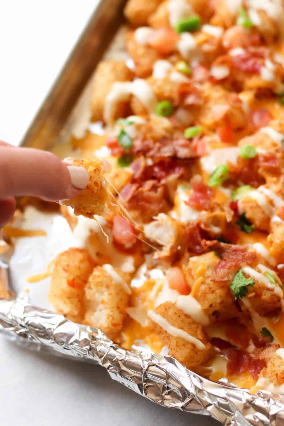 Hand picking up a cheesy tot from a baking sheet with loaded tater tots, bacon, cheese, and green onions on foil.