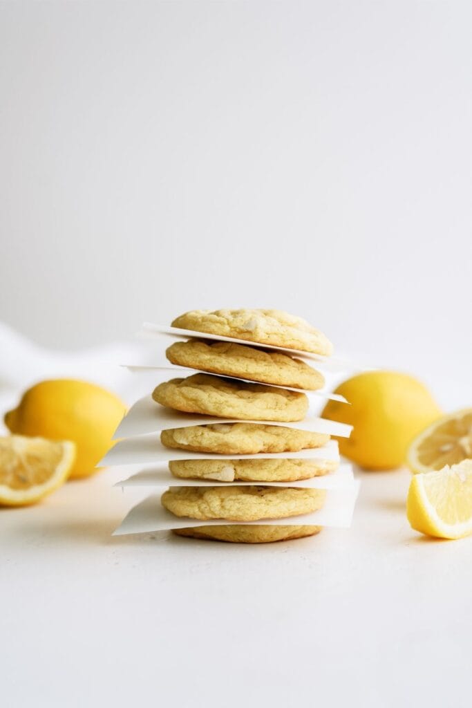 Lemon Cheesecake Pudding Cookies stacked with pieces of parchment in between each cookie and fresh lemons in the background.