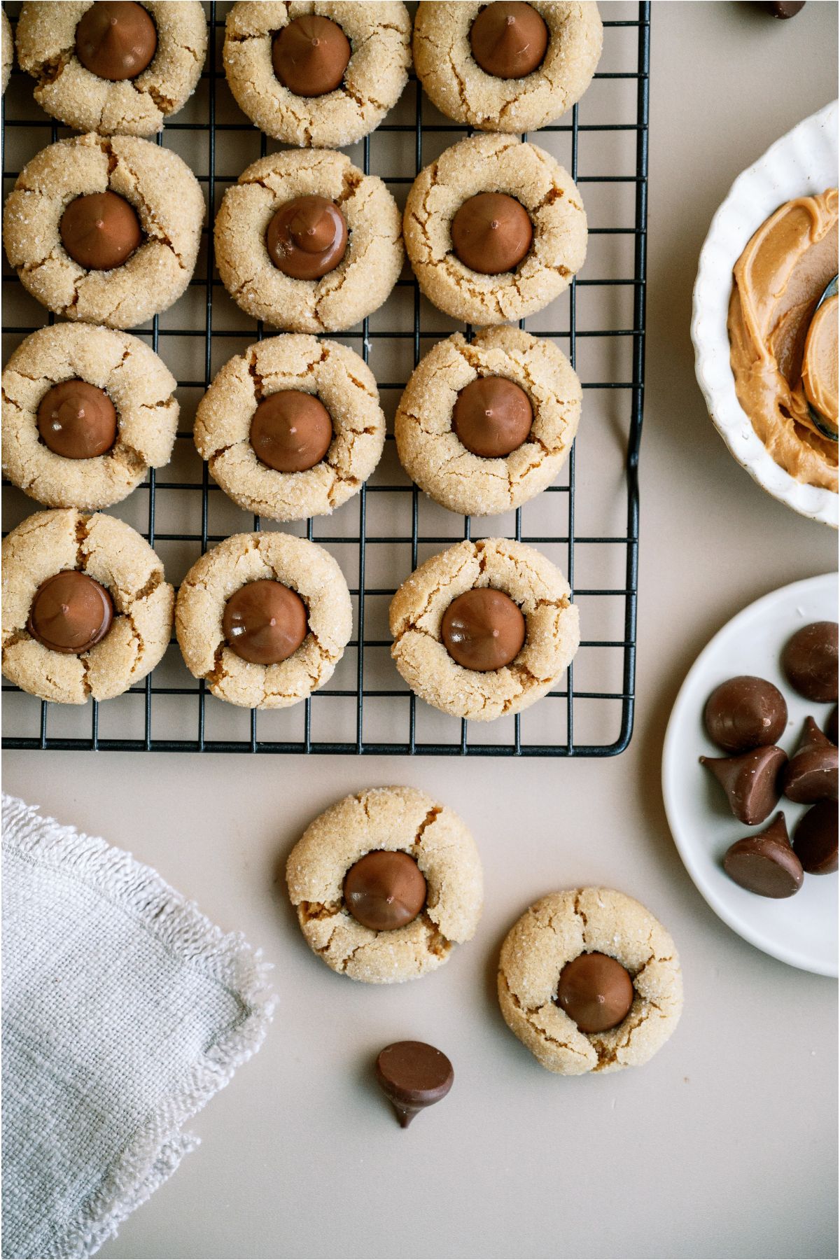 Top view of Peanut Butter Blossoms (Hershey’s Kiss Cookies) on a cooling rack, with two cookies on the counter. A bowl of chocolate kisses and a bowl of peanut butter off to the side.