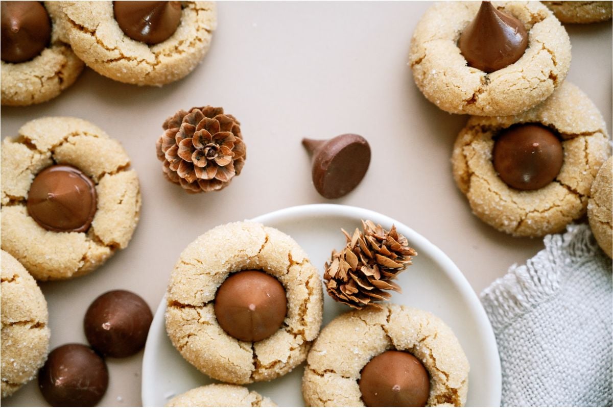 Peanut Butter Blossoms (Hershey’s Kiss Cookies) on a small plate with a small pine cone. Other Peanut Butter Blossoms (Hershey’s Kiss Cookies) around the plate.