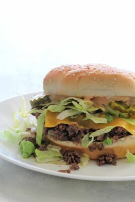 A sesame seed bun burger with lettuce, cheese, ground beef, and sauce sits on a white plate.