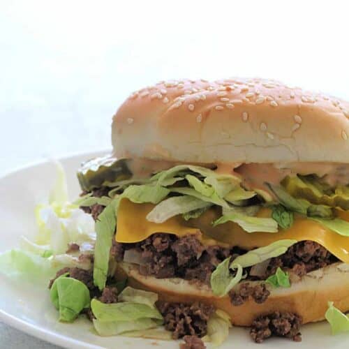 A sesame seed bun burger with lettuce, cheese, ground beef, and sauce sits on a white plate.