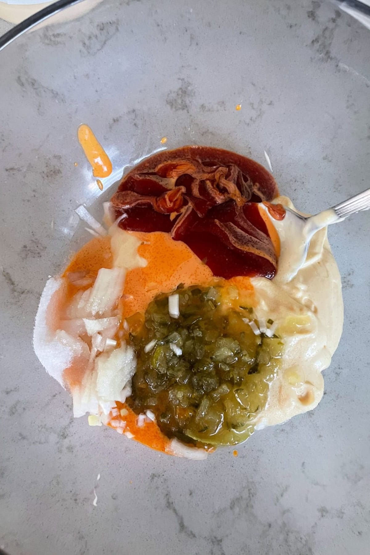 A mixing bowl containing various ingredients including diced onions, diced jalapeños, ketchup, bacon bits, and a sauce base. A spoon is in the bowl, ready for mixing.