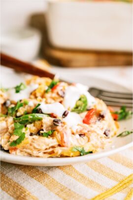 A close-up of a serving of Chicken Taco Casserole on a plate, showing melted cheese, diced tomatoes, chicken and chopped green onions. Topped with sour cream with a fork on the side.