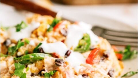 A close-up of a serving of Chicken Taco Casserole on a plate, showing melted cheese, diced tomatoes, chicken and chopped green onions. Topped with sour cream with a fork on the side.