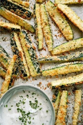 Crispy zucchini fries with breadcrumbs on parchment paper, accompanied by a bowl of creamy dipping sauce topped with chopped herbs.