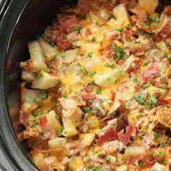 A slow cooker filled with a dish of diced potatoes, melted cheese, chopped bacon, and green onions.