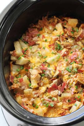 A slow cooker filled with a dish of diced potatoes, melted cheese, chopped bacon, and green onions.