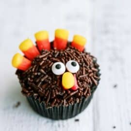 A cupcake decorated as a turkey with candy corn feathers, chocolate sprinkles, and candy eyes on a white surface. Text above reads: "10 of the EASIEST Thanksgiving Desserts" and "SIXSISTERSTUFF.COM".