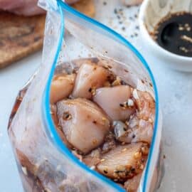 A plastic bag filled with marinated chicken pieces, placed on a countertop. A bowl with dark liquid and spices is in the background.