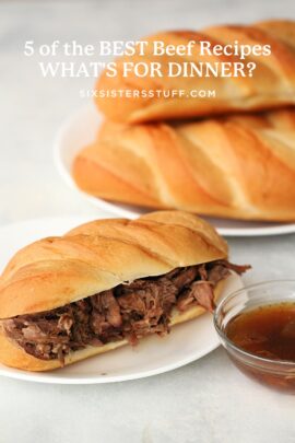 A sandwich with shredded beef and a side of dipping sauce is placed on a white plate, with other loaves of bread in the background.