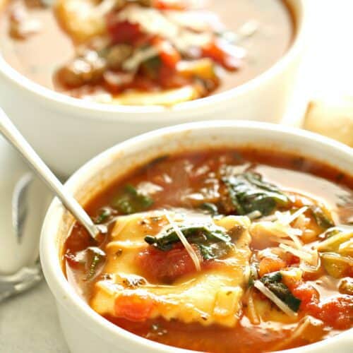 Two bowls of hearty soup featuring ravioli, tomatoes, and spinach, with a spoon in the front bowl.