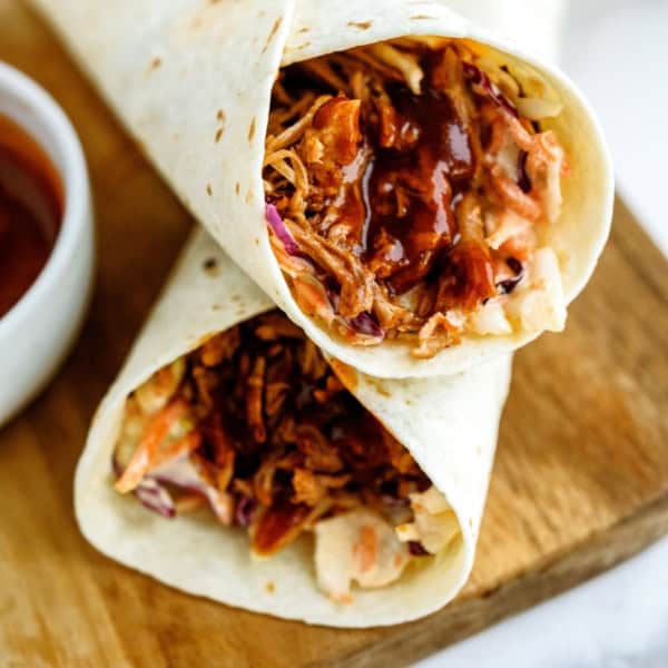 Close-up of two BBQ chicken wraps with shredded chicken, coleslaw, and barbecue sauce on a wooden surface. A small bowl of additional barbecue sauce is partially visible to the side.