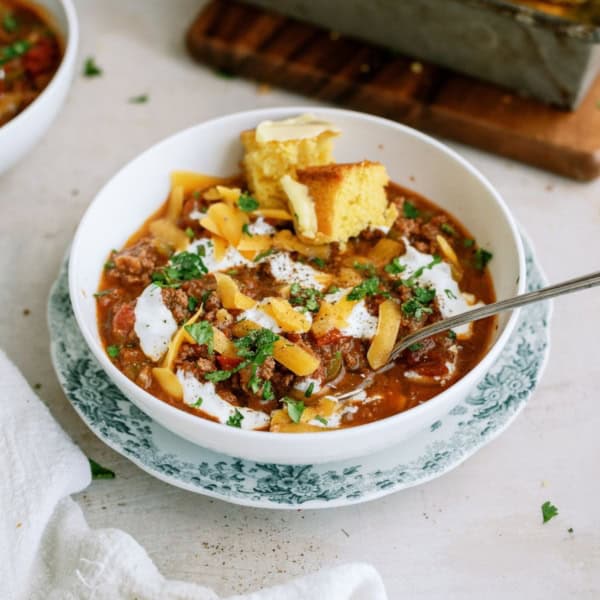 A bowl of chili topped with shredded cheese, sour cream, and chopped herbs, accompanied by cornbread on a floral-patterned plate.