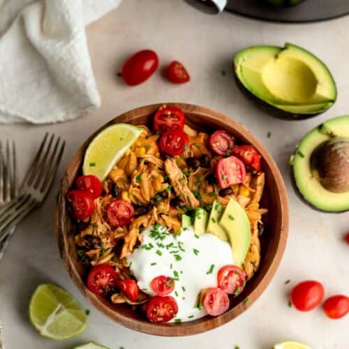 A wooden bowl filled with shredded chicken, avocado slices, cherry tomatoes, lime wedge, and a dollop of sour cream, garnished with chopped herbs. Forks, avocados, tomatoes, and lime pieces surround it.