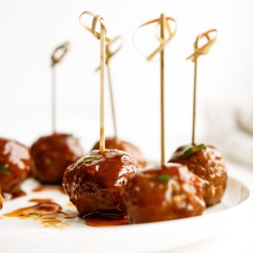 Meatballs glazed with sauce, each skewered with a decorative toothpick, are arranged on a white plate.