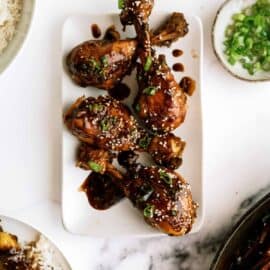 A rectangular plate with glazed chicken drumsticks garnished with sesame seeds and herbs. A small bowl of white rice and a small bowl of chopped green onions are visible on the side.