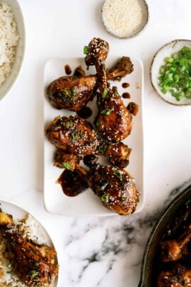 A rectangular plate with glazed chicken drumsticks garnished with sesame seeds and herbs. A small bowl of white rice and a small bowl of chopped green onions are visible on the side.