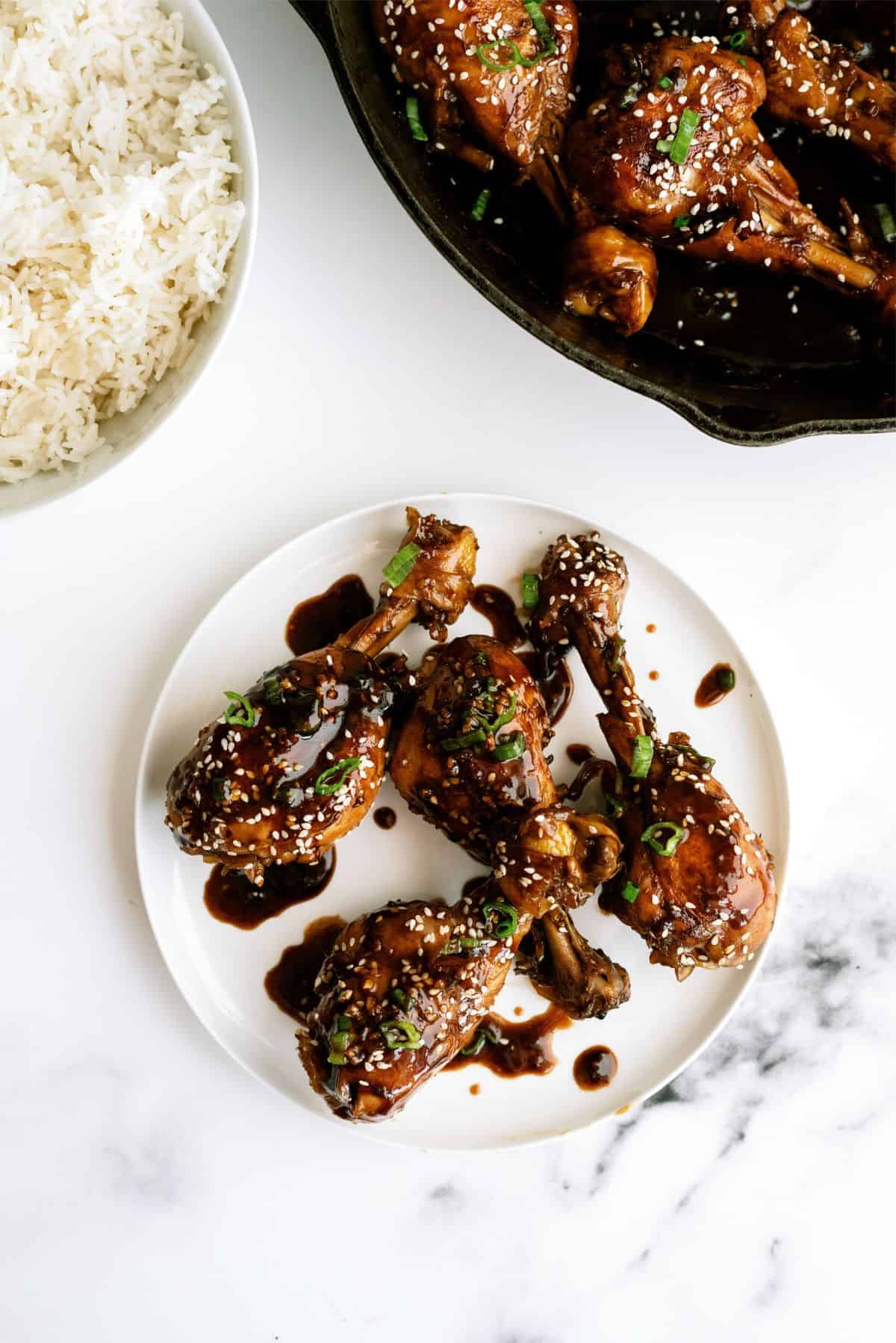 A plate of glazed chicken drumsticks garnished with sesame seeds and green onions, served next to a bowl of white rice.