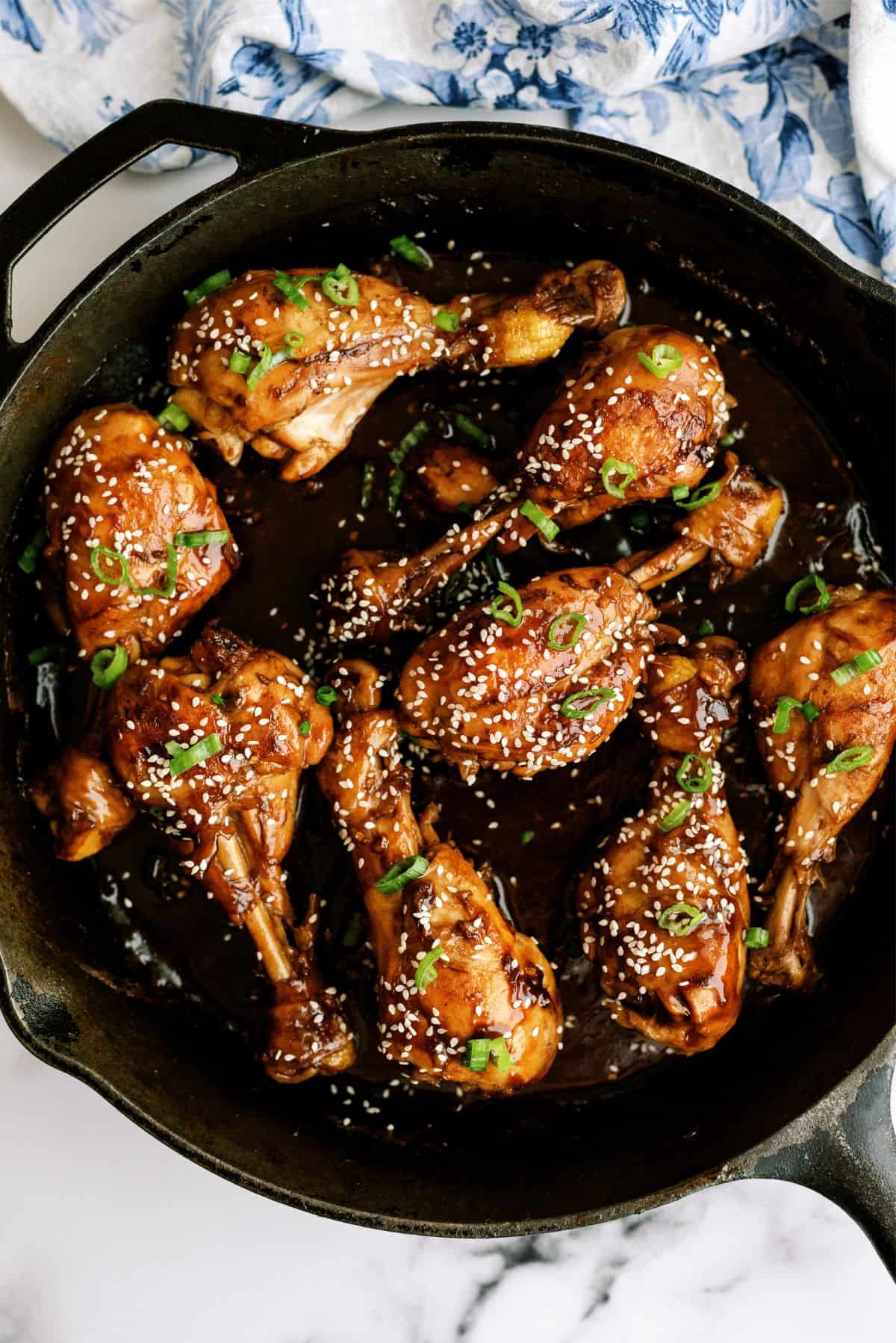 A skillet filled with glazed, cooked chicken drumsticks garnished with sesame seeds and chopped green onions.