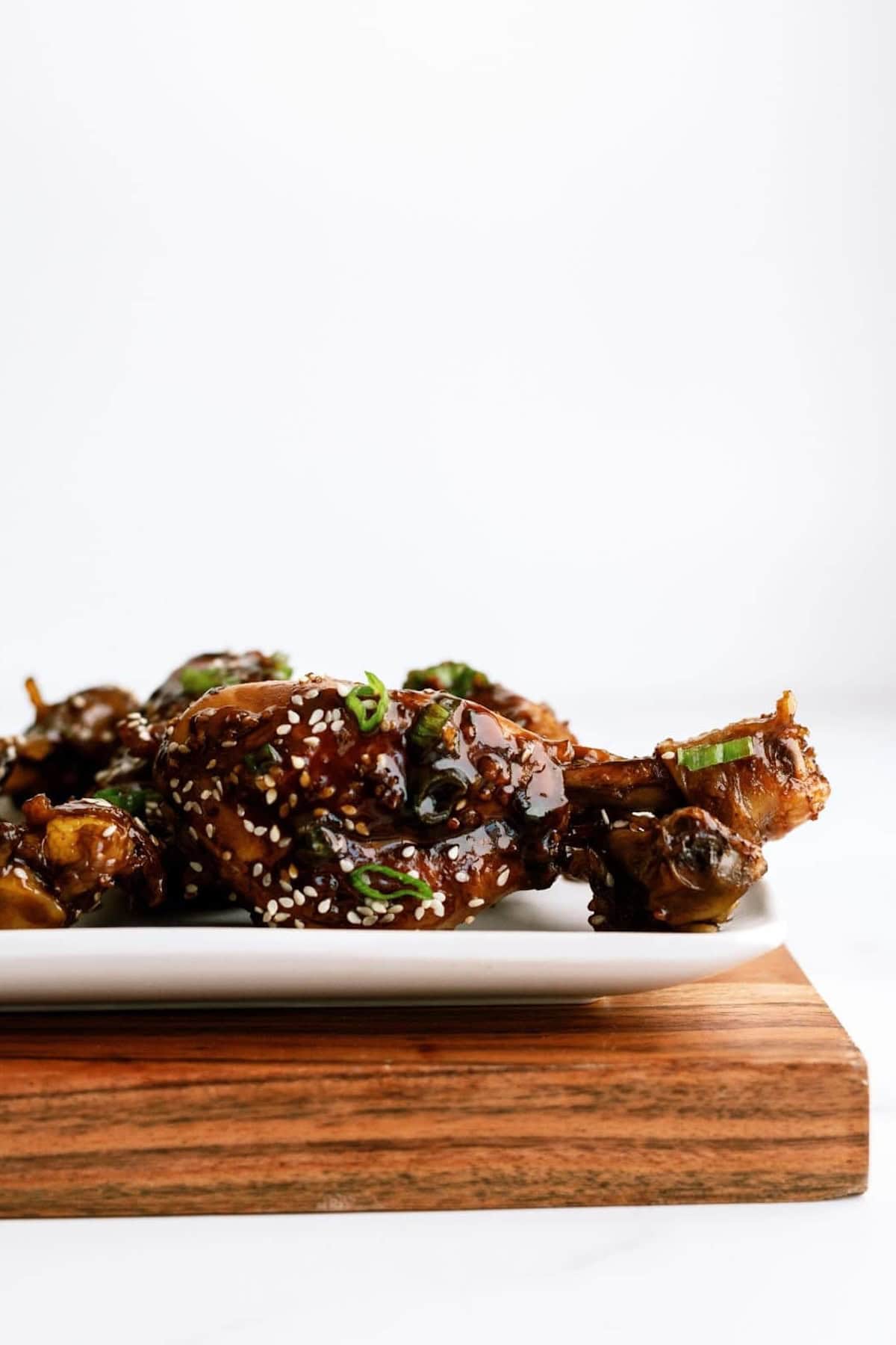 Plate of glazed chicken wings garnished with sesame seeds and chopped green onions, placed on a wooden board against a plain white background.