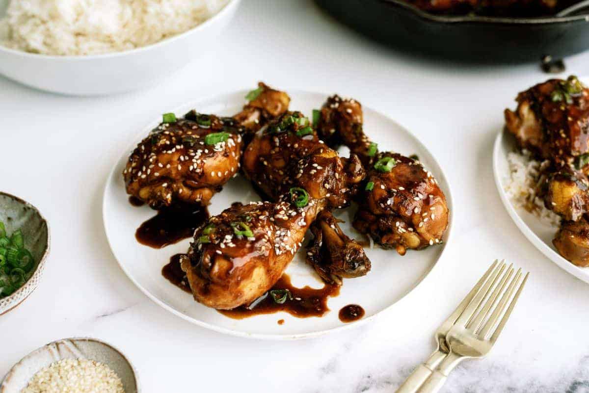 A plate of glazed chicken drumsticks garnished with sesame seeds and chopped green onions. A fork is placed beside the plate, and a bowl of white rice is in the background.