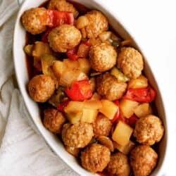 A white dish filled with meatballs, chunks of pineapple, red and green bell peppers, and a sauce, placed on a white cloth.