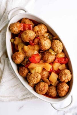 A white dish filled with meatballs, chunks of pineapple, red and green bell peppers, and a sauce, placed on a white cloth.