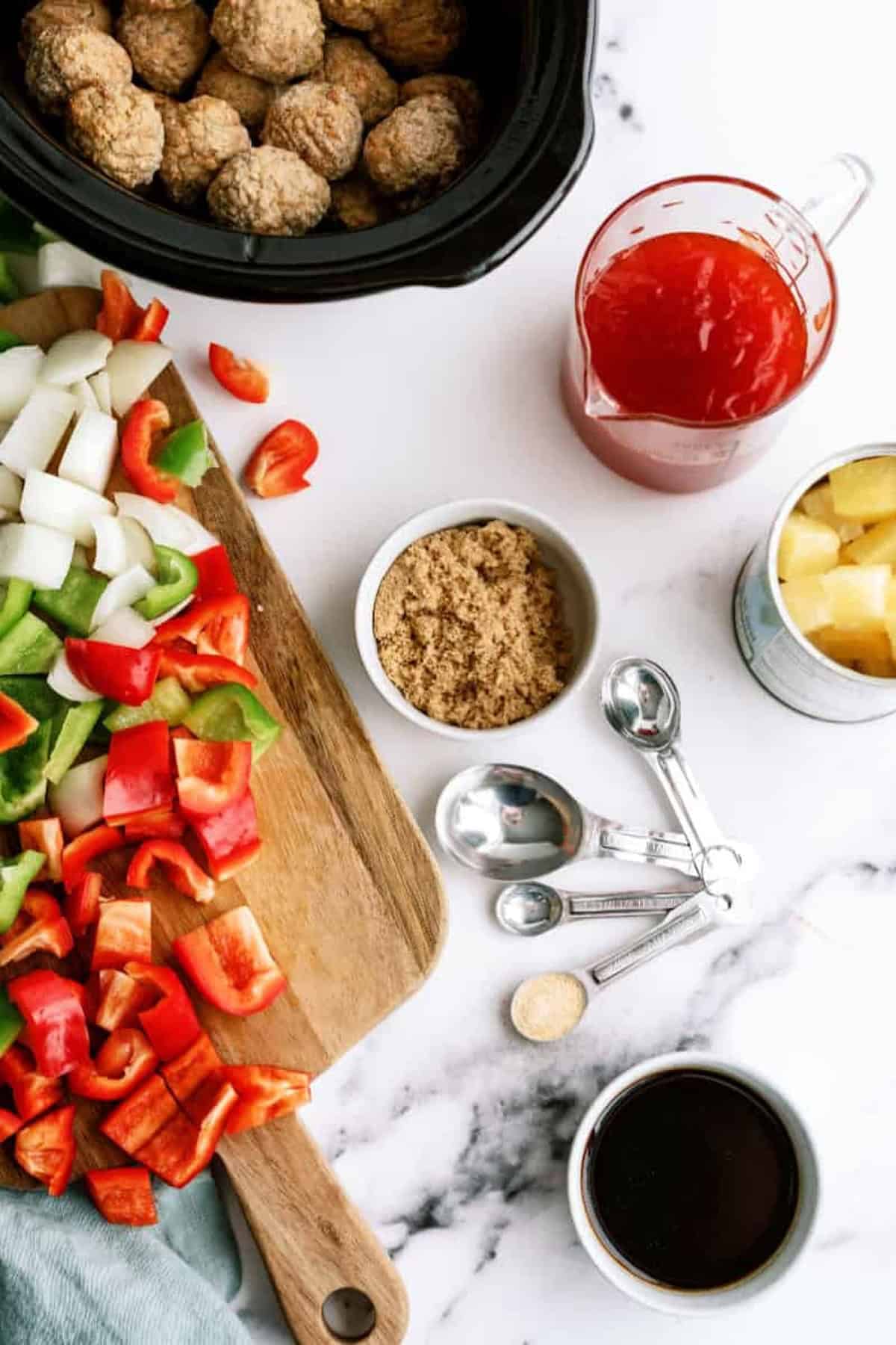 A crockpot with meatballs, a wooden board with chopped bell peppers and onions, a bowl of brown sugar, a cup of soy sauce, a measuring cup of red liquid, canned pineapple chunks, and measuring spoons.