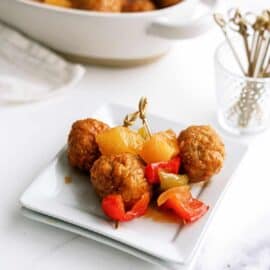 Meatballs with pineapple chunks and red bell peppers on a white plate, with a dish of more meatballs and a small container of toothpicks in the background.