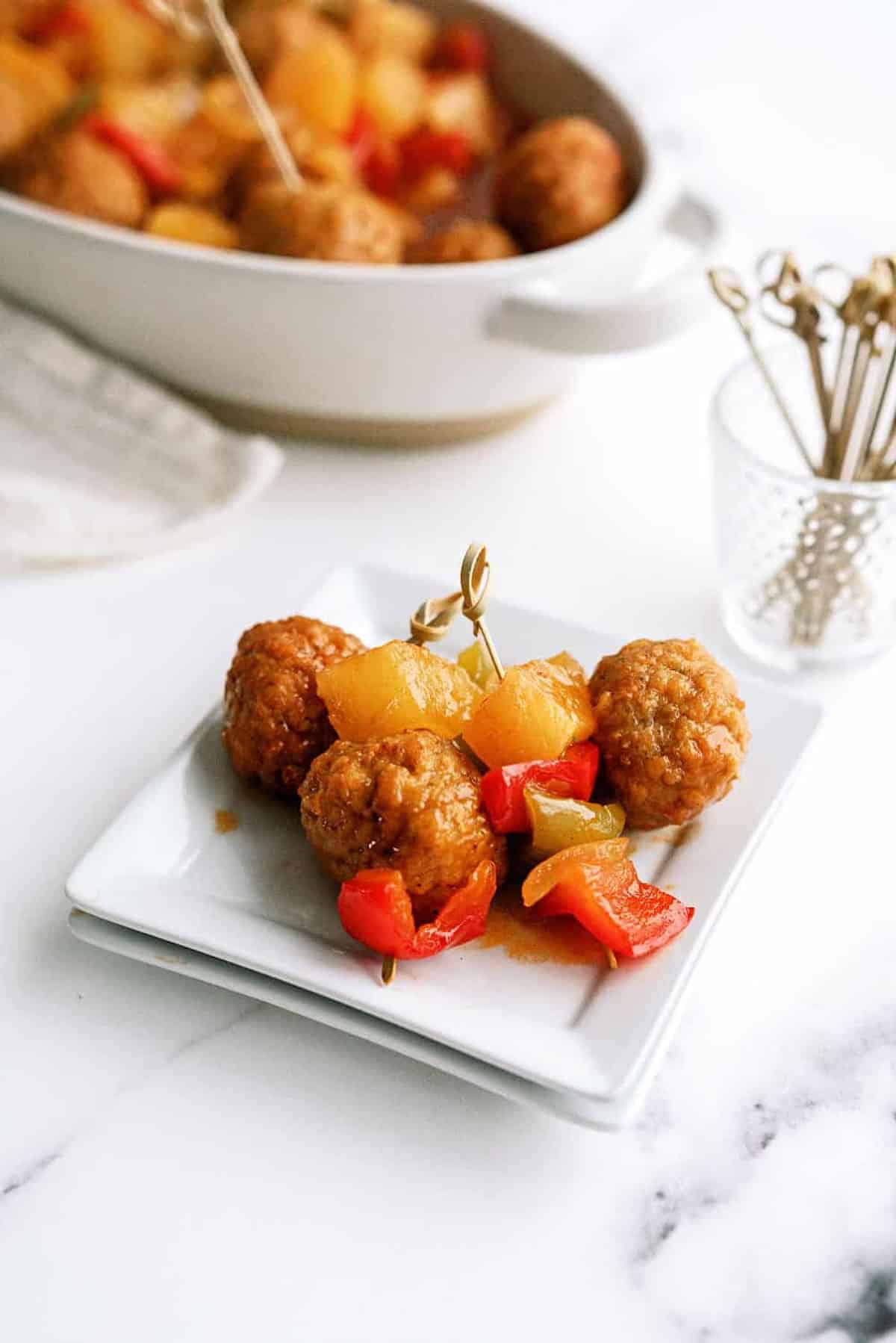 Meatballs with pineapple chunks and red bell peppers on a white plate, with a dish of more meatballs and a small container of toothpicks in the background.