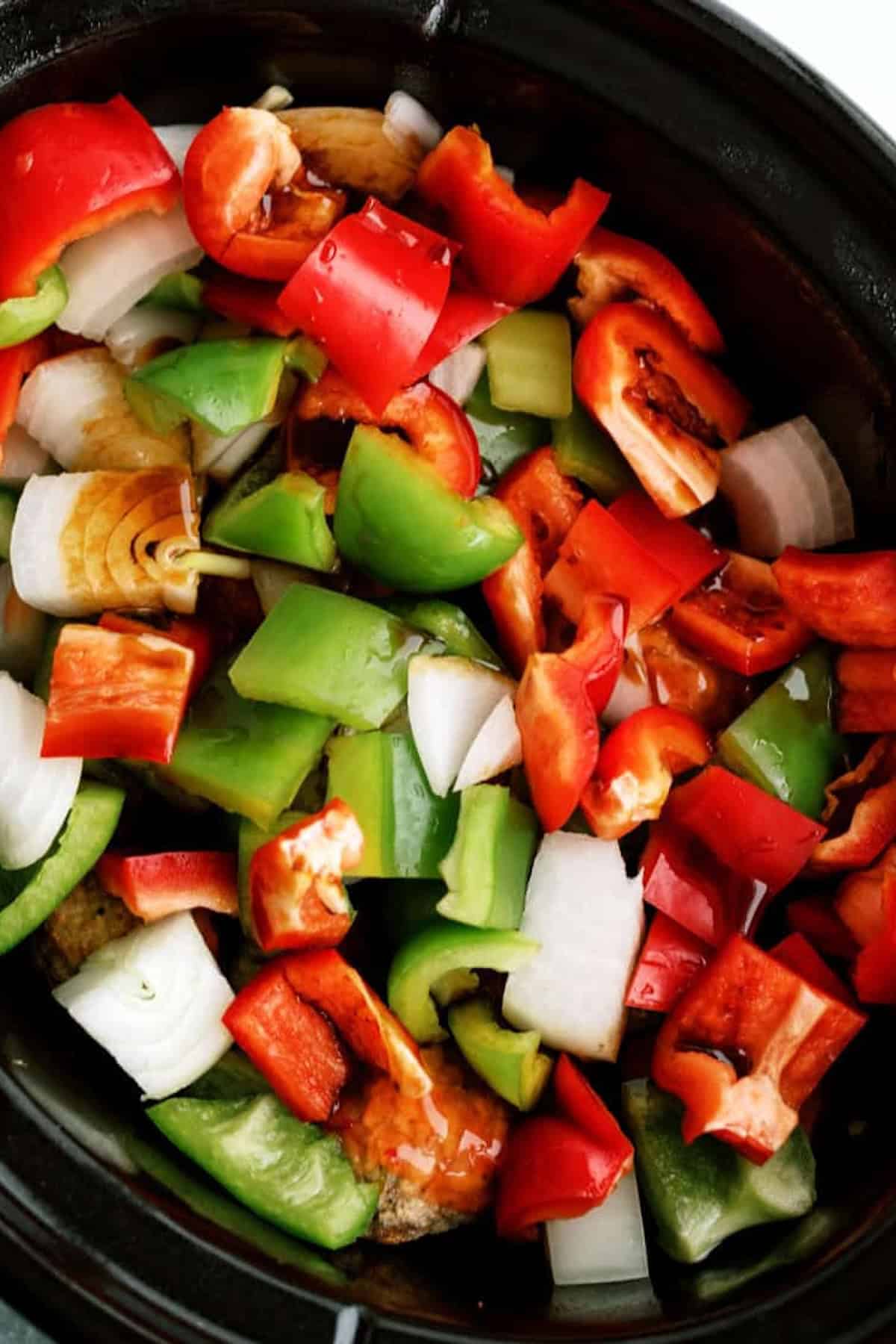 A slow cooker filled with chopped red and green bell peppers, onions, and garlic cloves, ready to be cooked.