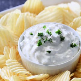 A bowl of creamy dip garnished with chopped herbs, surrounded by ridged potato chips.