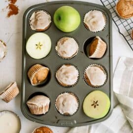 A muffin tin filled with apple muffins, some frosted and some plain. Green apple slices, a green apple, and a few muffins are scattered around. A spice jar and a cloth are present on the table.