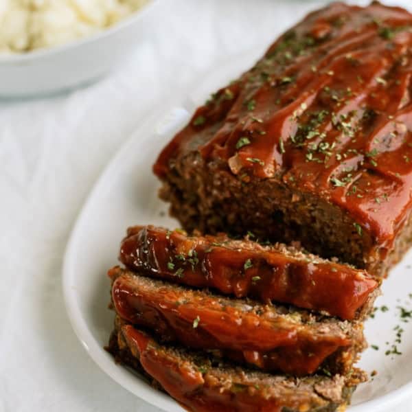 A meatloaf topped with a glaze, partially sliced, on a white plate. A bowl of mashed potatoes is visible in the background.