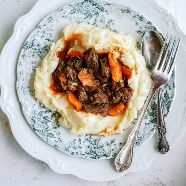 A plate with mashed potatoes topped with beef stew and carrots, served with a spoon and fork.